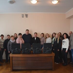 Групове фото працівників у приміщенні. Посередині стоїть стіл, на якому розміщено кілька чорних кейсів. Люди стоять півколом позаду столу, дивляться в об’єктив камери. Приміщення світле, з бежевими стінами та дерев’яною підлогою.