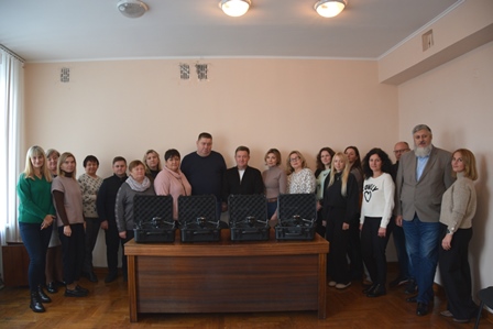 Групове фото працівників у приміщенні. Посередині стоїть стіл, на якому розміщено кілька чорних кейсів. Люди стоять півколом позаду столу, дивляться в об’єктив камери. Приміщення світле, з бежевими стінами та дерев’яною підлогою.