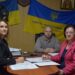 На фото троє працівників у кабінеті за круглим столом під час робочої наради. На стіні позаду розміщено два прапори України, один із гаслом «Єдність – наша головна сила». Люди мають перед собою документи та ручки, атмосфера ділова.