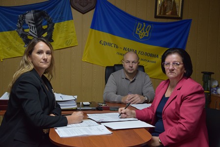 На фото троє працівників у кабінеті за круглим столом під час робочої наради. На стіні позаду розміщено два прапори України, один із гаслом «Єдність – наша головна сила». Люди мають перед собою документи та ручки, атмосфера ділова.