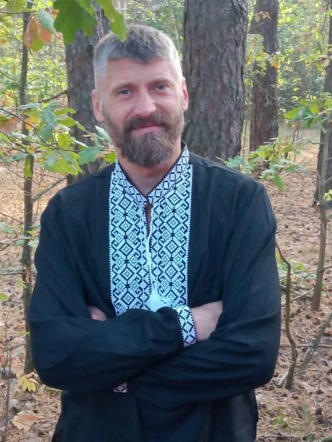 Чоловік стоїть у лісі серед дерев із опалим листям. На ньому чорна вишиванка з білим орнаментом. Він має коротке волосся, бороду й усміхається, схрестивши руки на грудях.