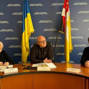 На фото зображено робочу нараду в адміністративному приміщенні. Троє посадовців сидять за круглим столом: двоє чоловіків і жінка. Вони ведуть обговорення та роблять записи.