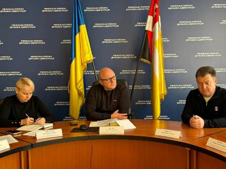 На фото зображено робочу нараду в адміністративному приміщенні. Троє посадовців сидять за круглим столом: двоє чоловіків і жінка. Вони ведуть обговорення та роблять записи.