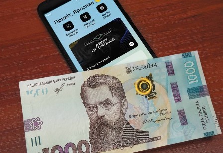 На столі лежить смартфон з відкритим додатком, на екрані якого видно привітальне повідомлення та банер “Army of Drones”. Перед смартфоном розміщена банкнота номіналом 1000 гривень