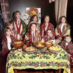 Група дітей і підлітків у традиційних українських вишиванках та барвистих квітчастих хустках стоїть і сидить навколо столу, застеленого яскравою вишитою скатертиною.