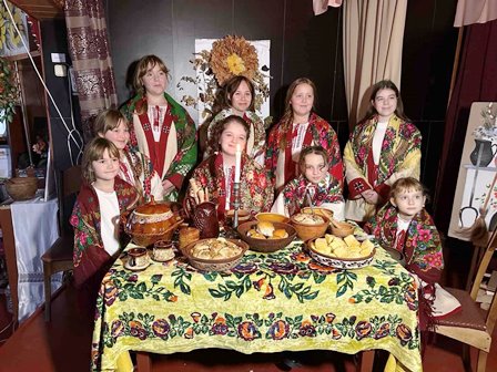 Група дітей і підлітків у традиційних українських вишиванках та барвистих квітчастих хустках стоїть і сидить навколо столу, застеленого яскравою вишитою скатертиною.