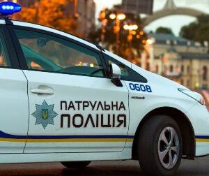 На фото службовий автомобіль патрульної поліції України з увімкненими проблисковими маячками. Машина білого кольору з синьо-жовтими смугами та написом «Патрульна поліція». На задньому плані — міська вулиця з будівлями та аркою.