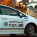 На фото службовий автомобіль патрульної поліції України з увімкненими проблисковими маячками. Машина білого кольору з синьо-жовтими смугами та написом «Патрульна поліція». На задньому плані — міська вулиця з будівлями та аркою.