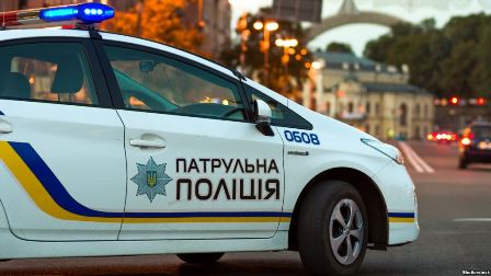 На фото службовий автомобіль патрульної поліції України з увімкненими проблисковими маячками. Машина білого кольору з синьо-жовтими смугами та написом «Патрульна поліція». На задньому плані — міська вулиця з будівлями та аркою.