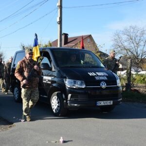 Колона українських військових іде сільською дорогою. Попереду рухається чорний мікроавтобус Volkswagen із написом «НА ВІЧНУ ПАМ’ЯТЬ ГЕРОЮ». Перед автомобілем несе прапор України військовослужбовець у камуфляжній формі.