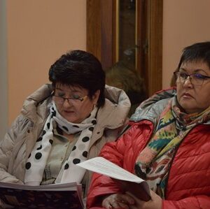 «Дві жінки сидять у приміщенні та уважно переглядають документи. Обидві в теплих куртках і шарфах; одна тримає в руках папку, інша — аркуші з паперами. На тлі видно дерев’яні двері та світлу стіну.»