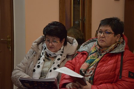 «Дві жінки сидять у приміщенні та уважно переглядають документи. Обидві в теплих куртках і шарфах; одна тримає в руках папку, інша — аркуші з паперами. На тлі видно дерев’яні двері та світлу стіну.»