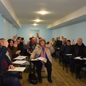 У світлій кімнаті з блакитними стінами сидить група людей за столами у два ряди. Вони тримають у руках документи та піднімають руки, голосуючи. Попереду в центрі сидить жінка в світлій куртці з шарфом, поруч – інші учасники засідання.