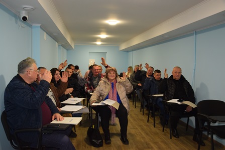 У світлій кімнаті з блакитними стінами сидить група людей за столами у два ряди. Вони тримають у руках документи та піднімають руки, голосуючи. Попереду в центрі сидить жінка в світлій куртці з шарфом, поруч – інші учасники засідання.