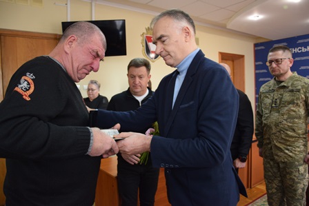 У приміщенні під час офіційної церемонії чоловік у темному светрі, помітно зворушений і засмучений, отримує з рук посадовця коробку з державною нагородою. Посадовець у темно-синьому костюмі та сорочці вручисто передає нагороду, тримаючи також букет квітів.