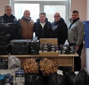 П’ятеро чоловіків стоять у приміщенні за великим столом, на якому розташовані коробки з дронами, комплекти Starlink, генератор, екофлу та мішки з маскувальними сітками. Усі посміхаються та дивляться в камеру, демонструючи передану допомогу для військових