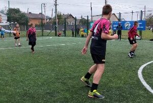 На фото зображено групу підлітків, які грають у футбол на штучному полі просто неба. Один гравець у яскравій помаранчево-чорній формі готується зробити передачу, а кілька інших — у рожево-чорних і бордових футболках — уважно стежать за м’ячем.