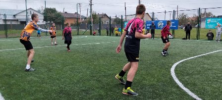 На фото зображено групу підлітків, які грають у футбол на штучному полі просто неба. Один гравець у яскравій помаранчево-чорній формі готується зробити передачу, а кілька інших — у рожево-чорних і бордових футболках — уважно стежать за м’ячем.