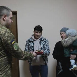 У приміщенні чоловік у військовій формі простягає нагороду двом жінкам. Одна з них тримає червону коробку з відзнакою, інша — маленьку дитину на руках. Усі стоять поруч, атмосфера зосереджена та урочиста.