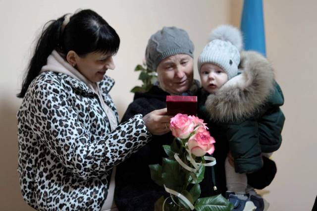 Три особи стоять разом у приміщенні: жінка у светрі з леопардовим принтом показує невелику подарункову коробочку літній жінці в сірій шапці, яка тримає на руках маленьку дитину в теплому комбінезоні з хутряним капюшоном.