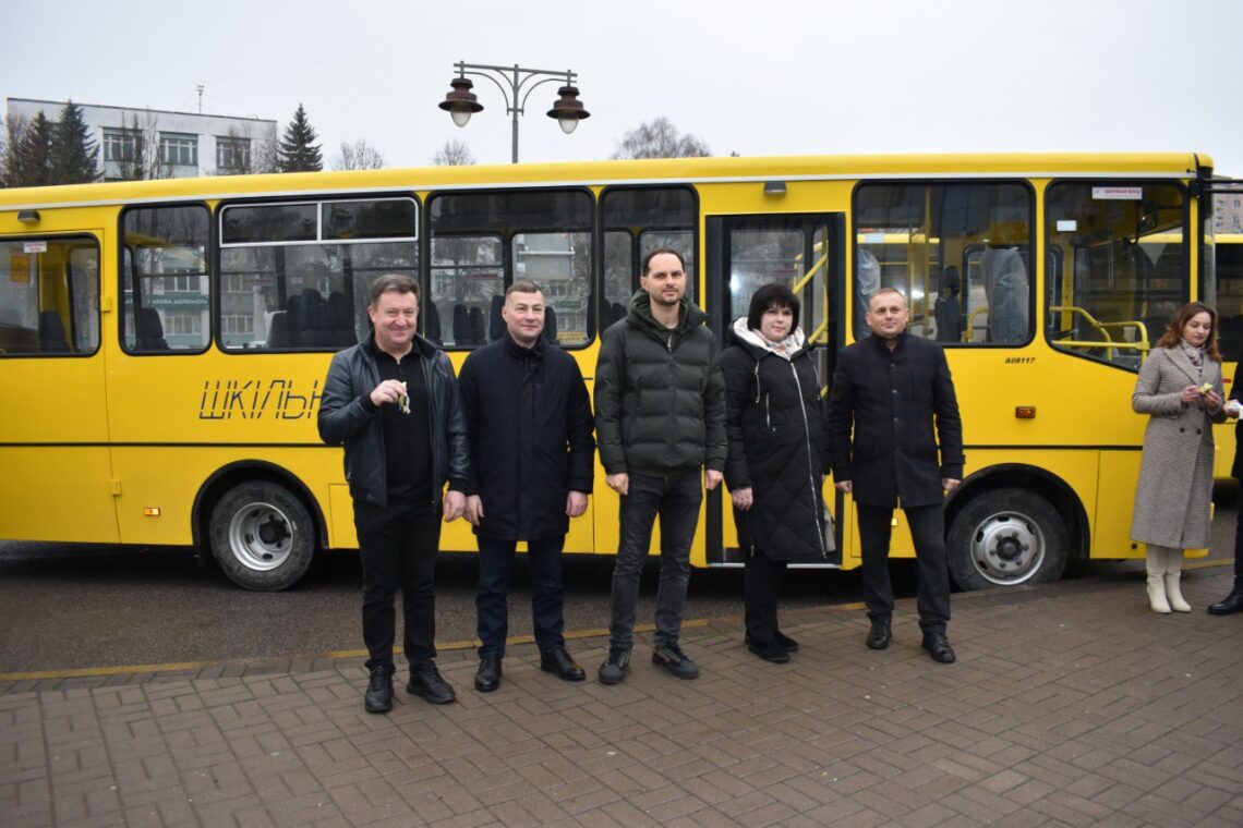 На фото зображено групу людей, які стоять перед жовтим шкільним автобусом. Один із чоловіків тримає в руках ключі від транспорту. Усі учасники події позують для фото на тлі автобуса з відчиненими дверима.