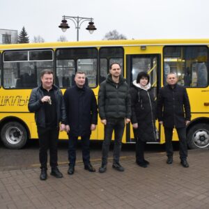На фото зображено групу людей, які стоять перед жовтим шкільним автобусом. Один із чоловіків тримає в руках ключі від транспорту. Усі учасники події позують для фото на тлі автобуса з відчиненими дверима.