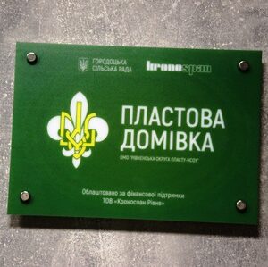На фото зелена інформаційна табличка на стіні. На ній зображено емблему Пласту та текст: «Пластова домівка», а також логотипи Городоцької сільської ради та компанії Kronospan. Табличка закріплена чотирма металевими кріпленнями на сіруватій фактурній поверхні.
