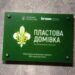 На фото зелена інформаційна табличка на стіні. На ній зображено емблему Пласту та текст: «Пластова домівка», а також логотипи Городоцької сільської ради та компанії Kronospan. Табличка закріплена чотирма металевими кріпленнями на сіруватій фактурній поверхні.