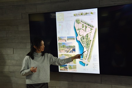 Студентка презентує проєкт благоустрою громадського простору, вказуючи на план території з зеленими зонами та водним об’єктом, відображений на великому екрані.