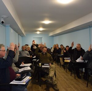 У приміщенні зали засідань відбувається сесія сільської ради: за партами сидять депутати, більшість з яких підняли руки під час голосування. На столах — документи та папки.