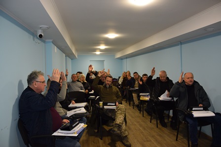 У приміщенні зали засідань відбувається сесія сільської ради: за партами сидять депутати, більшість з яких підняли руки під час голосування. На столах — документи та папки.