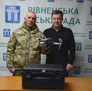 Двоє чоловіків стоять за столом у приміщенні з брендованим фоном «Рівненська міська рада». Один чоловік у військовій формі, поруч — чоловік у цивільному одязі. На столі перед ними розміщений чорний захисний кейс з обладнанням.