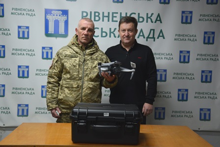 Двоє чоловіків стоять за столом у приміщенні з брендованим фоном «Рівненська міська рада». Один чоловік у військовій формі, поруч — чоловік у цивільному одязі. На столі перед ними розміщений чорний захисний кейс з обладнанням.