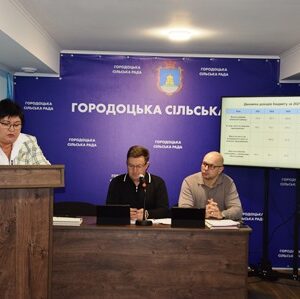 Позачергова сесія Городоцької сільської ради: у залі засідань за столом сидять сільський голова та депутати, ліворуч виступає доповідач за трибуною, на екрані — презентація з показниками бюджету громади.