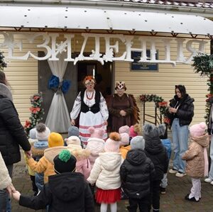 На фото група дітей та дорослих стоїть перед входом до будівлі, прикрашеної новорічними гірляндами. Над входом великими літерами розміщено напис «РЕЗИДЕНЦІЯ». Біля дверей стоять дві людини у святкових костюмах, які вітають маленьких відвідувачів.