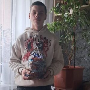 Молодий чоловік у світлому худі тримає в руках прозорий пакунок із солодощами. Він стоїть у приміщенні біля вікна та кімнатної рослини, дивиться в камеру.