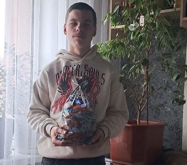 Молодий чоловік у світлому худі тримає в руках прозорий пакунок із солодощами. Він стоїть у приміщенні біля вікна та кімнатної рослини, дивиться в камеру.