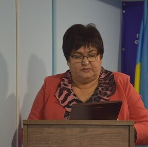 Жінка у червоному піджаку виступає за трибуною, читаючи текст з планшета; на тлі — прапор України та банер сільської ради.