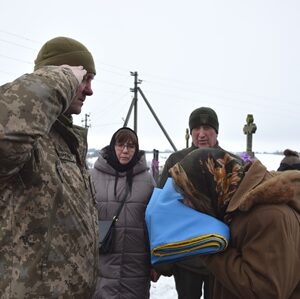 Військовослужбовець у зимовій формі віддає військове вітання під час церемонії прощання. Поруч жінка тримає складений Державний Прапор України, навколо — родичі, односельці та учасники жалобного заходу на кладовищі в зимову пору.