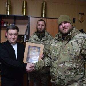 У службовому кабінеті троє чоловіків: двоє військовослужбовців у камуфляжній формі та представник органу місцевого самоврядування. Вони тиснуть руки, один із військових тримає подяку в рамці. На задньому плані — шафа з нагородами та символікою