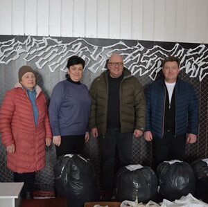 Група з п’яти людей стоїть у приміщенні перед декоративною сіткою; перед ними на підлозі розміщені пакети з гуманітарною допомогою та маскувальними матеріалами, підготовленими для потреб Збройних Сил України.