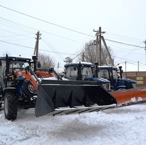Три комунальні трактори з відвалами для прибирання снігу, припарковані на засніженій території населеного пункту.