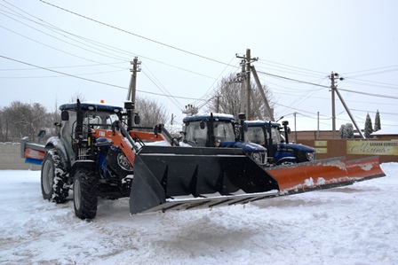 Три комунальні трактори з відвалами для прибирання снігу, припарковані на засніженій території населеного пункту.