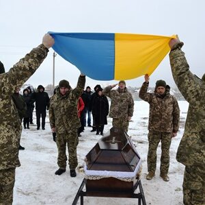 Військові Збройних сил України тримають розгорнутий синьо-жовтий державний прапор над труною загиблого воїна під час церемонії прощання. Поруч — побратими та жителі громади, які в скорботі віддають останню шану захиснику