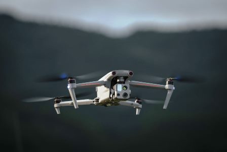 Квадрокоптерний дрон із камерою на підвісі летить у повітрі на тлі розмитих гір і неба, знятий крупним планом спереду