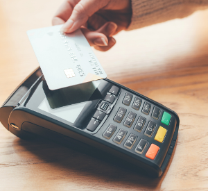 Рука тримає банківську картку та прикладає її до платіжного термінала для безконтактної оплати на дерев’яній поверхні.