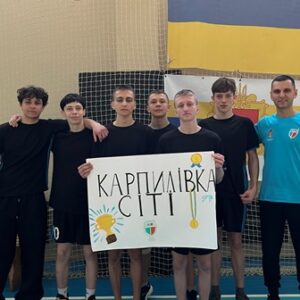 На фото — команда юнаків у спортивній формі разом із тренером у спортивному залі. Хлопці стоять пліч-о-пліч і тримають плакат з написом «Карпилівка Сіті», прикрашений малюнками кубка, медалі та емблеми