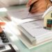 На столі лежать документи та калькулятор, поруч – макет будинку. Людина тримає ручку й працює з паперами, виконуючи фінансові розрахунки, ймовірно пов’язані з нерухомістю або оформленням житла.