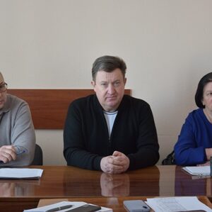 Троє людей сидять за дерев’яним столом у світлому приміщенні під час робочої наради. Перед ними лежать документи та канцелярське приладдя. Усі мають зосереджений вигляд і дивляться вперед, що створює ділову атмосферу обговорення