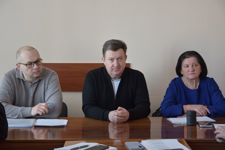 Троє людей сидять за дерев’яним столом у світлому приміщенні під час робочої наради. Перед ними лежать документи та канцелярське приладдя. Усі мають зосереджений вигляд і дивляться вперед, що створює ділову атмосферу обговорення